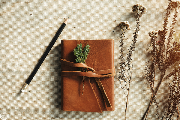 A leather journal tied with a sprig of greenery beside a pencil and dried flowers on linen
