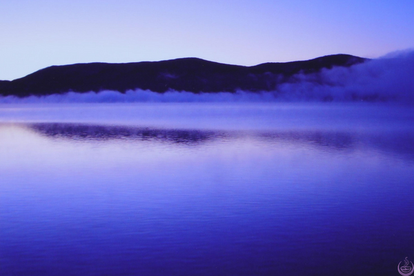 Mist drifting over a still lake at dawn with a dark ridge behind