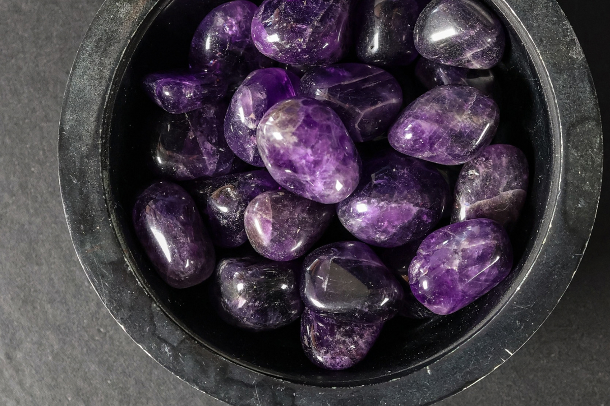A black bowl of amethyst tumbled stones on a dark surface