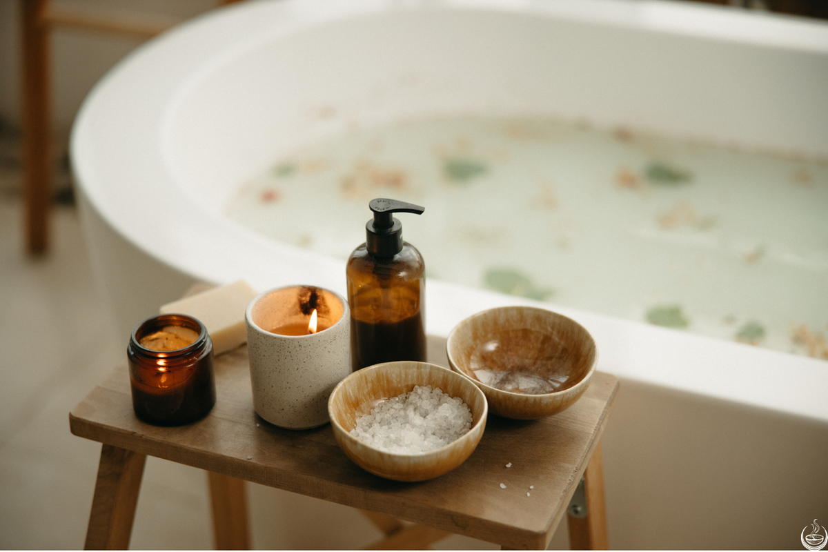A bowl of bath salt, a candle and an amber bottle on a wooden stool beside a drawn bath