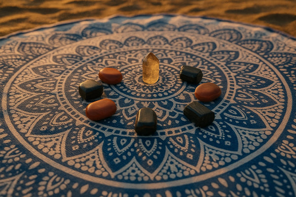 A circle of grounding crystals like Black Tourmaline, Red Jasper, and Smoky Quartz laid out on a beach tapestry during golden hour.