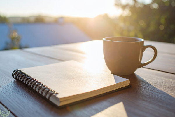 A closed spiral notebook and a mug on a wooden table at sunrise