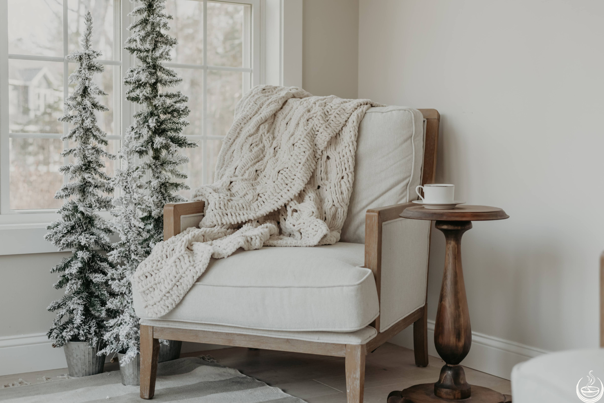 A cream armchair with a knitted blanket beside snow-dusted fir trees and a window