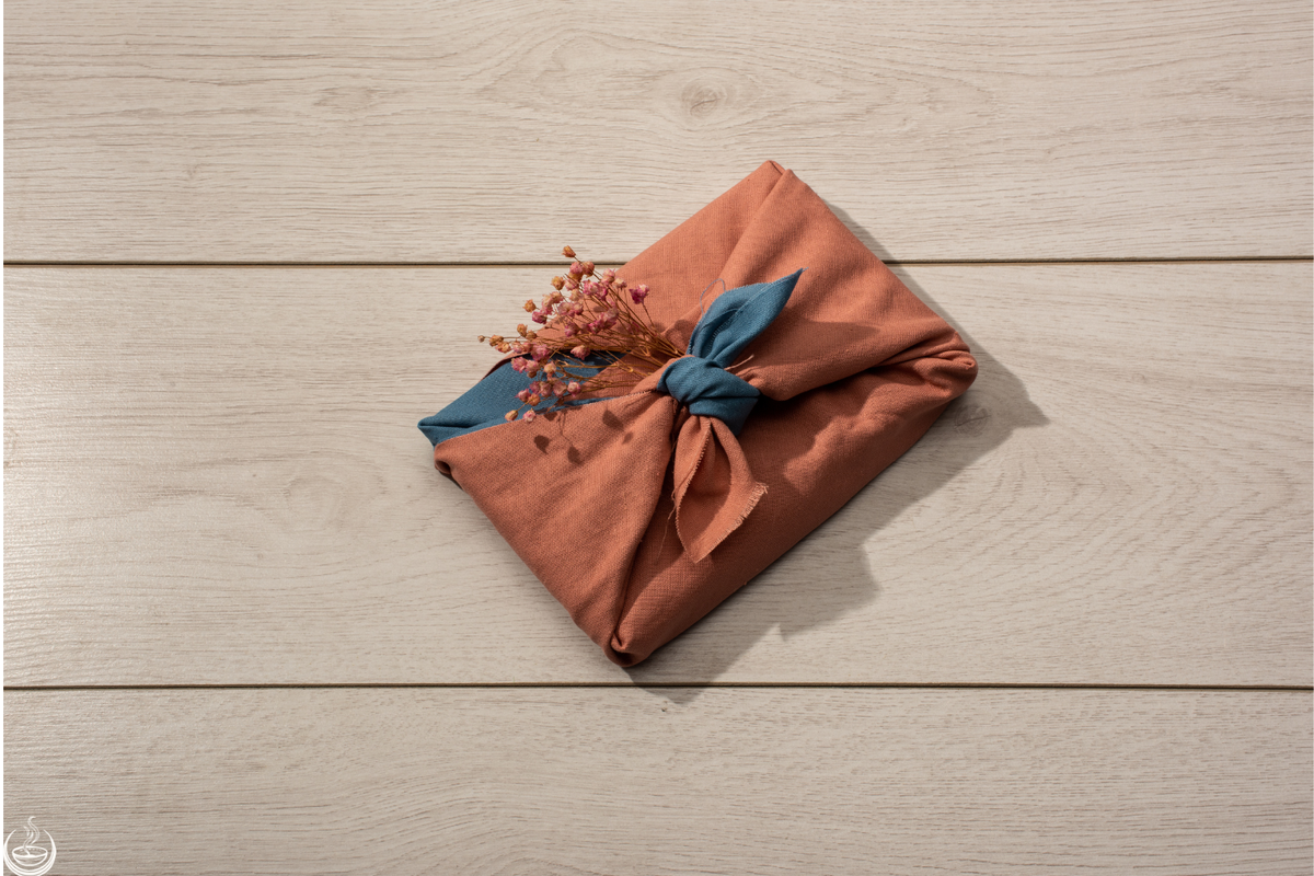 A gift wrapped in terracotta cloth and tied with a blue knot and dried flowers, on a pale wood table