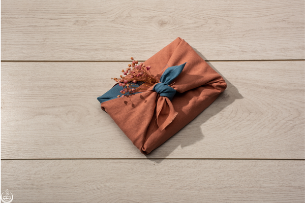 A gift wrapped in terracotta cloth and tied with a blue knot and dried flowers, on a pale wood table