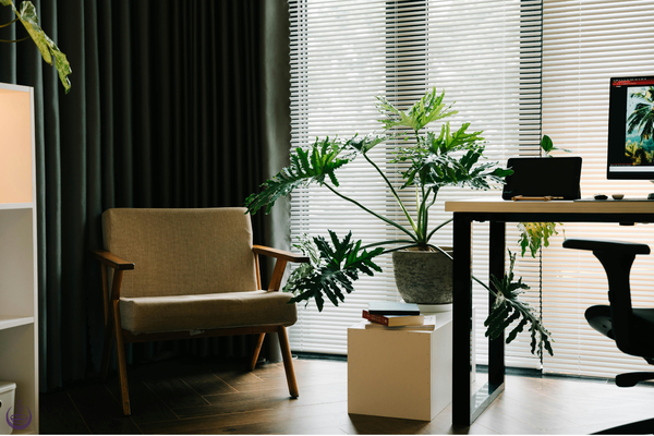 A home office with a desk, chair and large plant beside a window with blinds