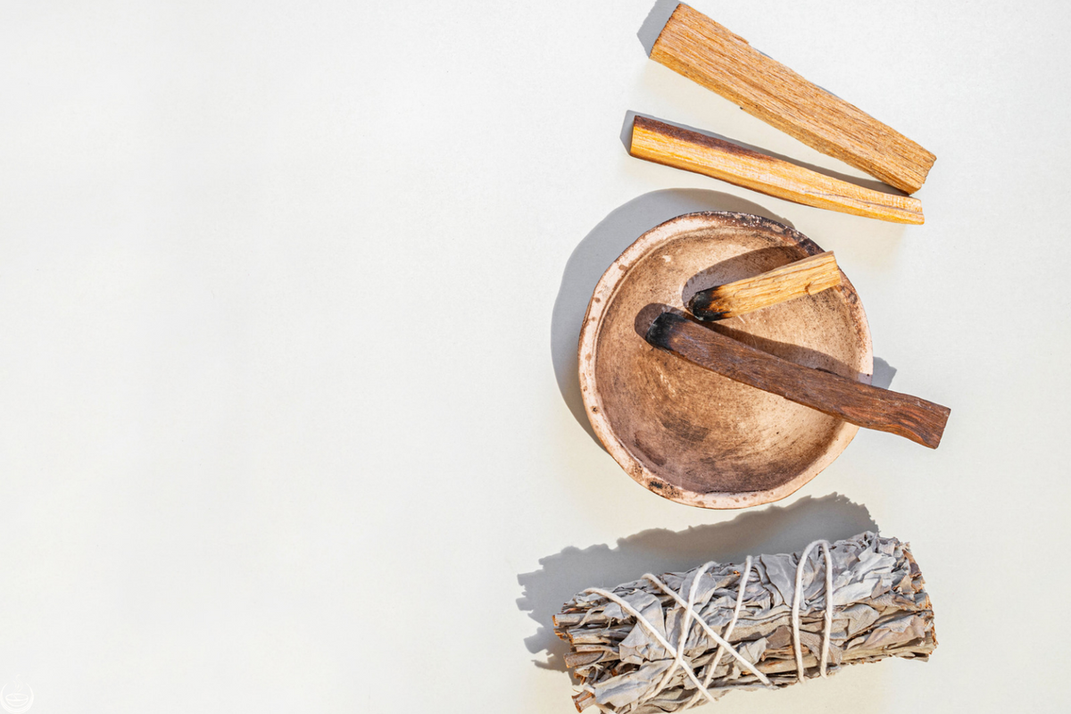A white sage bundle, palo santo sticks, and a burnt stick in a clay bowl, seen from above