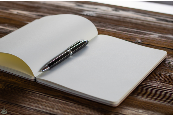 An open blank notebook with a black pen resting on it, on a dark wooden table
