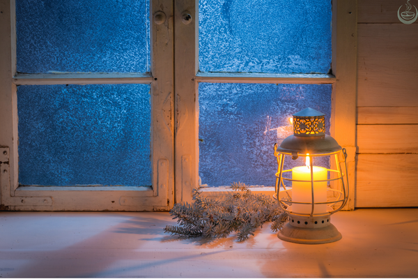 Candle glowing inside a lantern on a frosted windowsill in winter, with a sprig of fir beside it