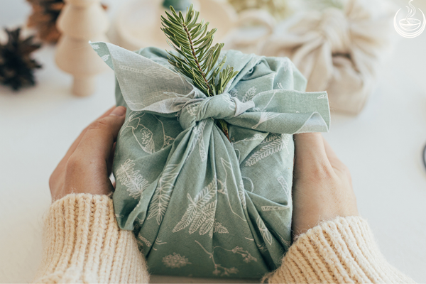 Hands holding a gift wrapped in sage green fabric and topped with an evergreen sprig