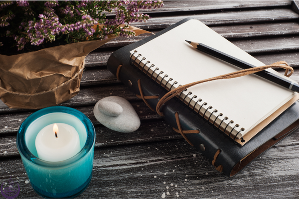 Lit candle, journal and pencil, and a smooth grey stone on a wooden table
