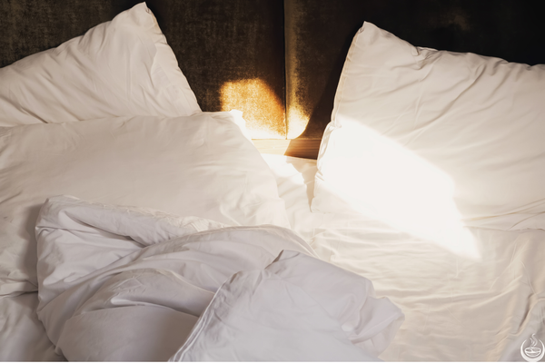 Unmade white bed with morning sunlight falling across the pillows