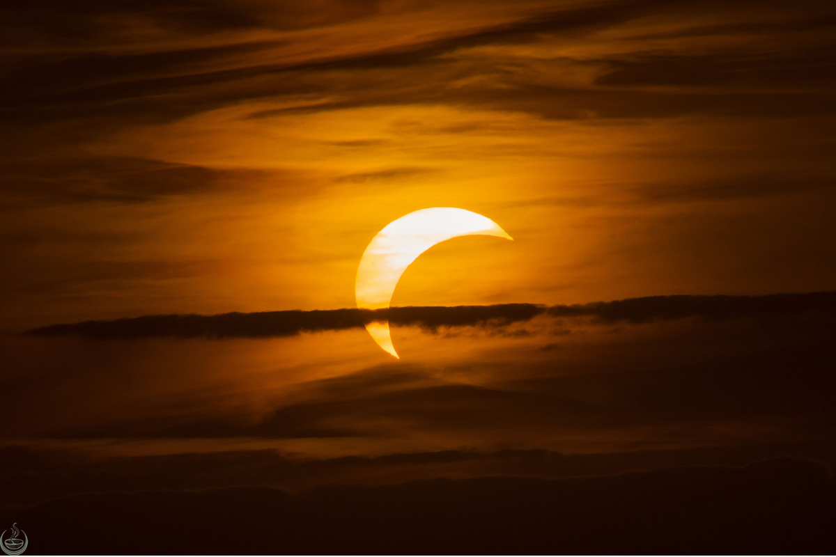 A partial solar eclipse glowing orange behind a band of cloud at sunset