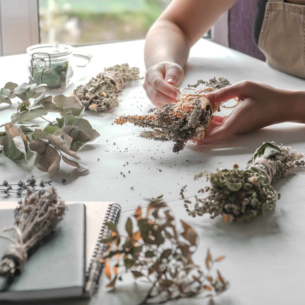 Hands arranging dried plants on a table with a notebook in the foreground.