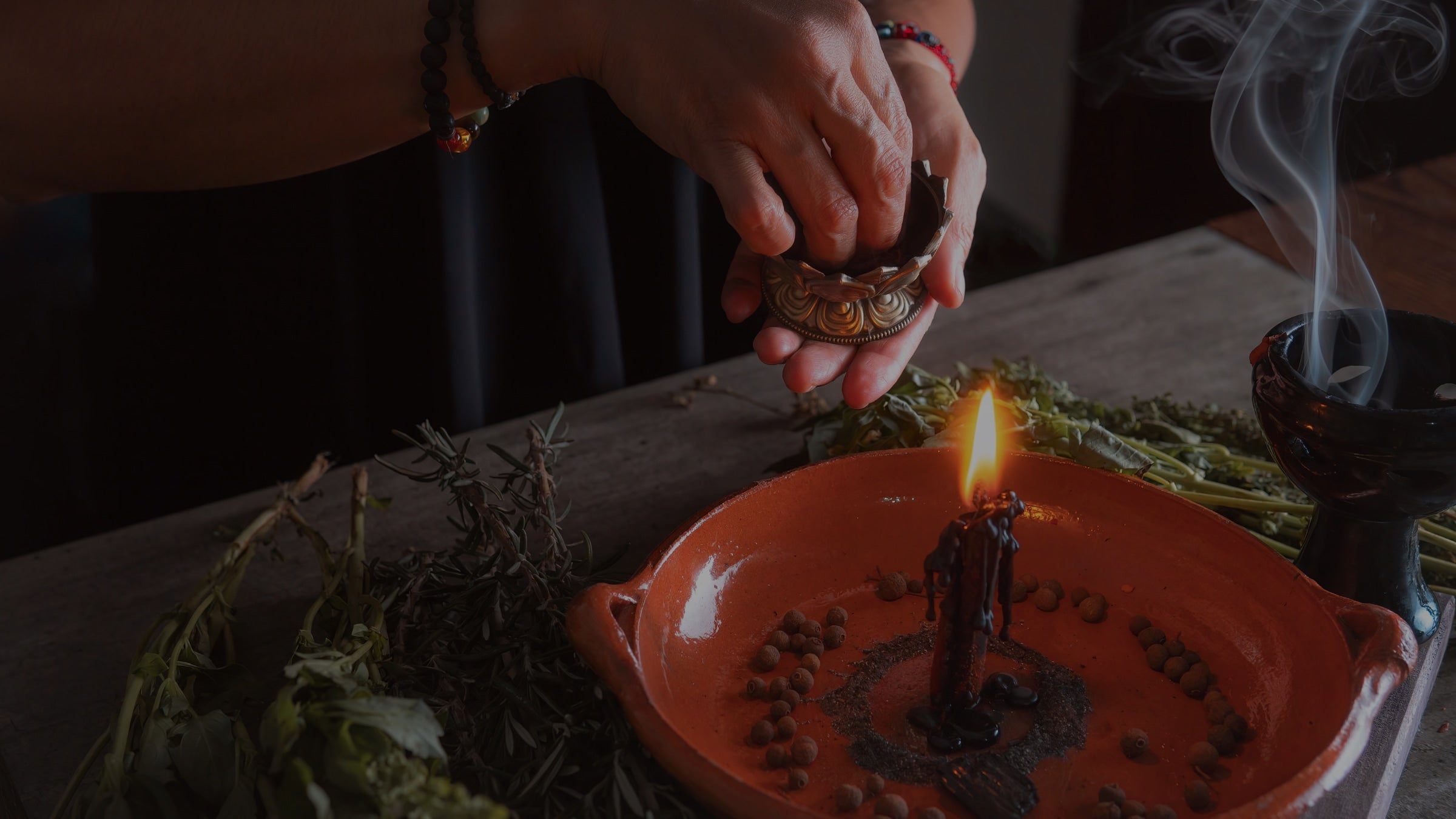 Hand lighting a candle with smoke rising, on a wooden surface with herbs.